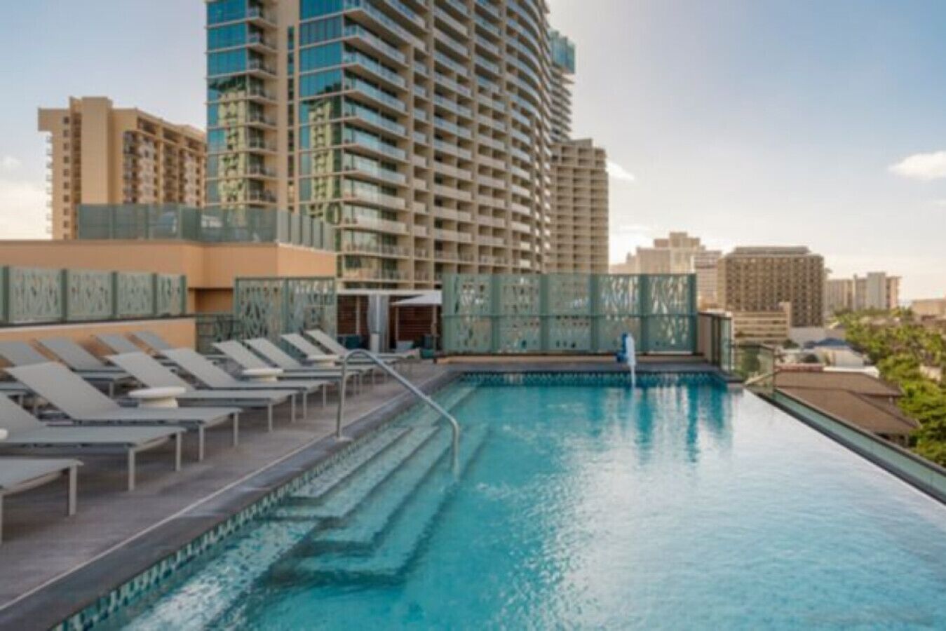 A rooftop pool with lounge chairs, a glass balustrade, and high-rise buildings in the background.