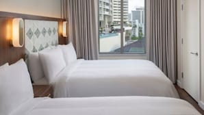 A hotel room with two beds, a headboard with light fixtures, a window with curtains, and a view of buildings and a pool. - Marriott Vacation Club, Waikiki (1BR) (Honolulu)