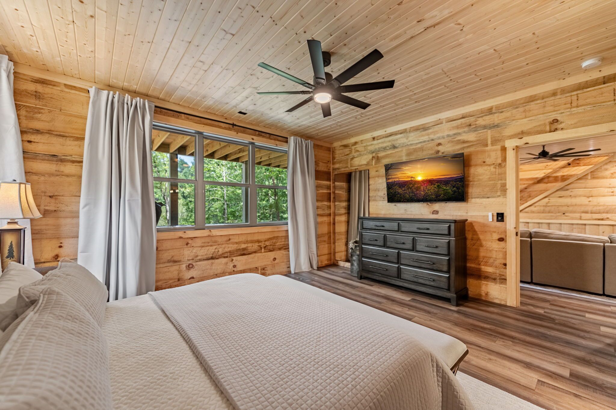 A wooden cabin bedroom with a bed, a dresser, a television, and a ceiling fan.