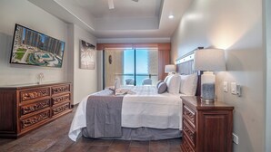 A hotel room with a large bed, a wooden dresser, a nightstand, a lamp, and a view of the outside.