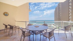 A balcony with wicker chairs and a round table, overlooking a beach and high-rise buildings.