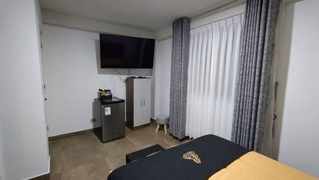 A hotel room with a bed, a flat-screen TV mounted on the wall, a small desk with a computer, and a window with curtains.