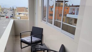 A balcony with two wicker chairs and a small table, overlooking a street with buildings and parked cars. - MAXIMUM COMFORT (Distrito de Chilca)