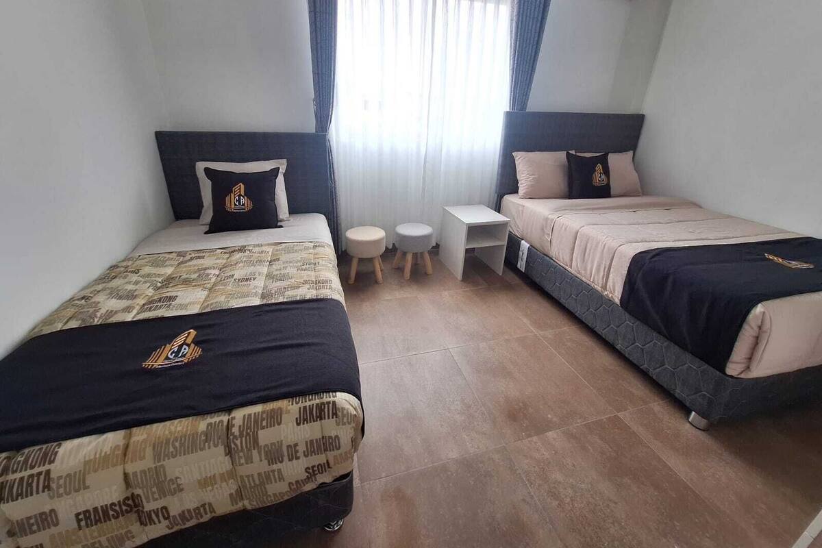 Quarto de hotel com duas camas, uma mesinha e uma janela com cortinas.