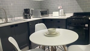 Eine moderne Küche mit grauen Fliesen an der Spritzschutzwand, einer Kaffeemaschine, einer Mikrowelle, einem Wasserkocher, einem Herd und einem Essbereich mit weißem Tisch und Stühlen.