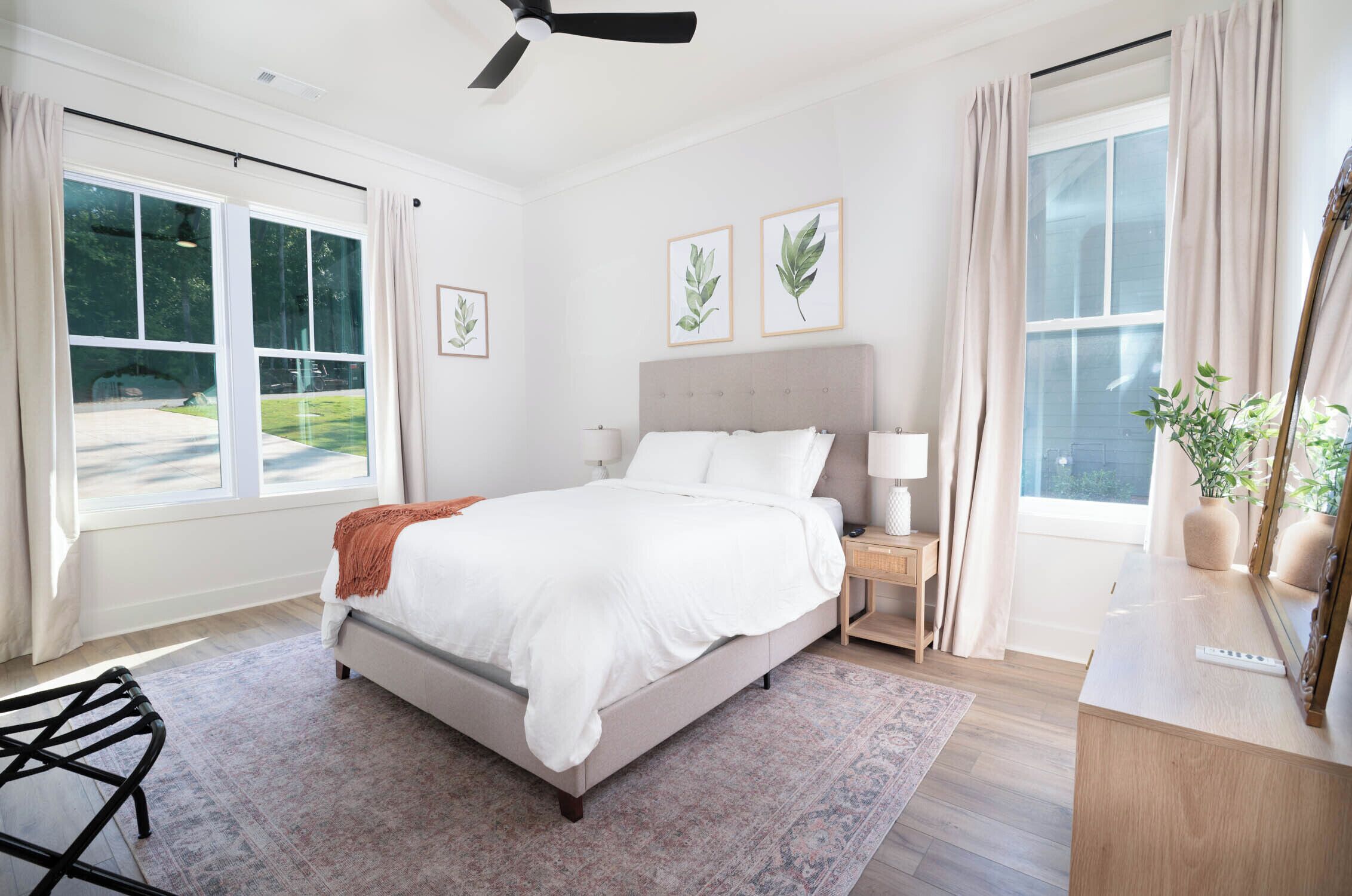 A bedroom with a bed, bedside tables, a ceiling fan, and large windows.