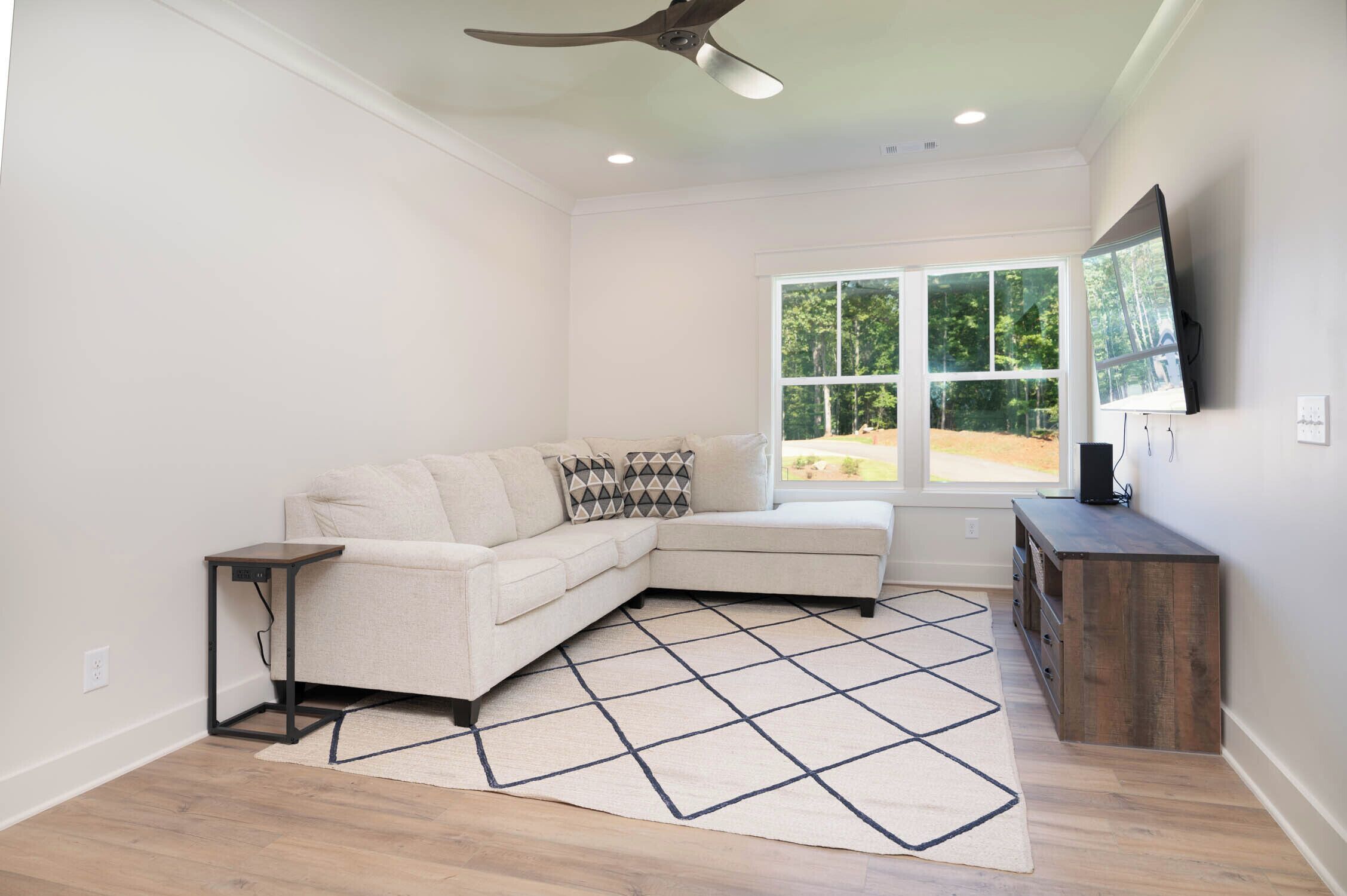 A living room with a sectional sofa, a small side table, a wooden cabinet, and a large window with a view of trees.