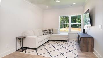A living room with a sectional sofa, a small side table, a wooden cabinet, and a large window with a view of trees.