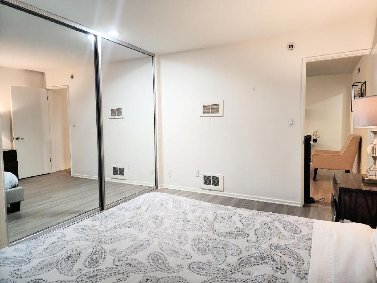 Une chambre avec un grand miroir, un lit recouvert d’une couvre-lit à motifs, une chaise et un bureau avec une lampe.