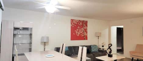 Una sala de estar moderna con comedor, un ventilador de techo, una obra de arte floral roja y una cocina al fondo.
