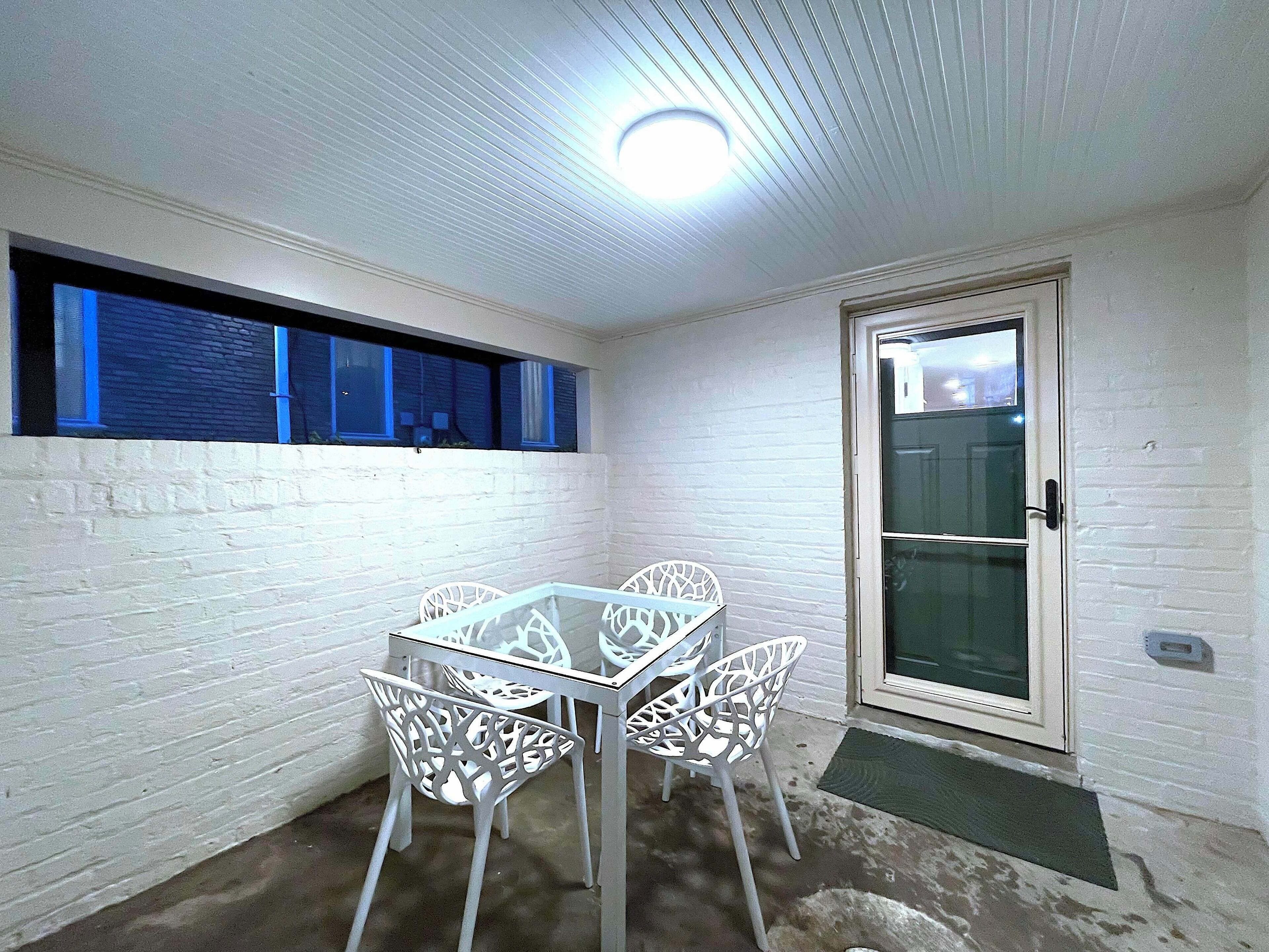 Un petit patio clos, doté d’une table à plateau de verre et de chaises blanches, d’une porte avec un panneau de verre et d’une fenêtre avec des volets bleus.