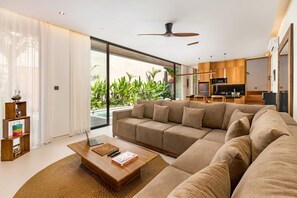 A modern living room with a large sectional sofa, a wooden coffee table, and a view of a pool and greenery outside.