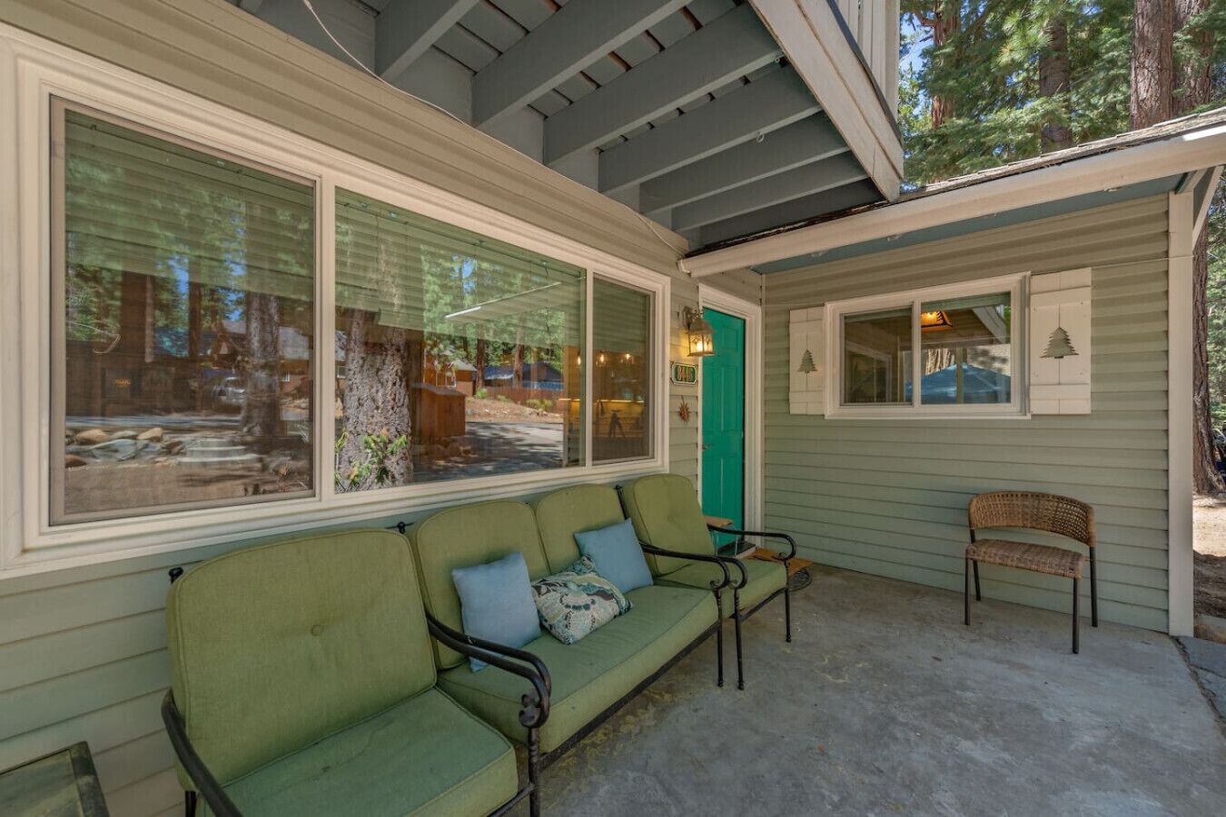 A covered porch with a green sofa and a wicker chair, large windows, and a small decorative tree.
