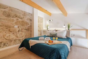 Ein gemütliches Schlafzimmer mit Steinwand, Holzbalkendecke, einem Bett mit türkisfarbener Bettdecke, zwei Nachttischlampen und einem Tablett mit Frühstücksangeboten.