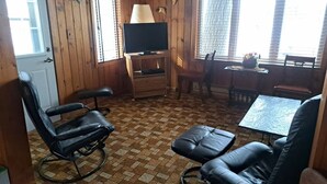 Une pièce aux murs lambrés, équipée d’un téléviseur, d’une table et de deux fauteuils à bascule.