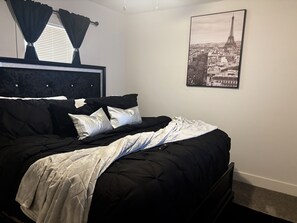 Un dormitorio con cama, ropa de cama blanco y negro, cabecero negro, ventana con cortinas negras y un cuadro enmarcado de la Torre Eiffel en la pared.