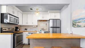 A modern kitchen with white cabinets, stainless steel appliances, a wooden island with stools, and a herringbone-patterned backsplash. - Sinatra Cottage · Classic Charm by Ocean Town (Galveston)