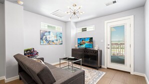 A living room with a gray sofa, a glass coffee table, a TV on a black cabinet, and artwork on the walls. - Sinatra Cottage · Classic Charm by Ocean Town (Galveston)