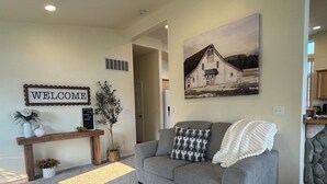 A living room with a gray sofa, a welcome sign, a painting of a barn, and a stone accent wall.