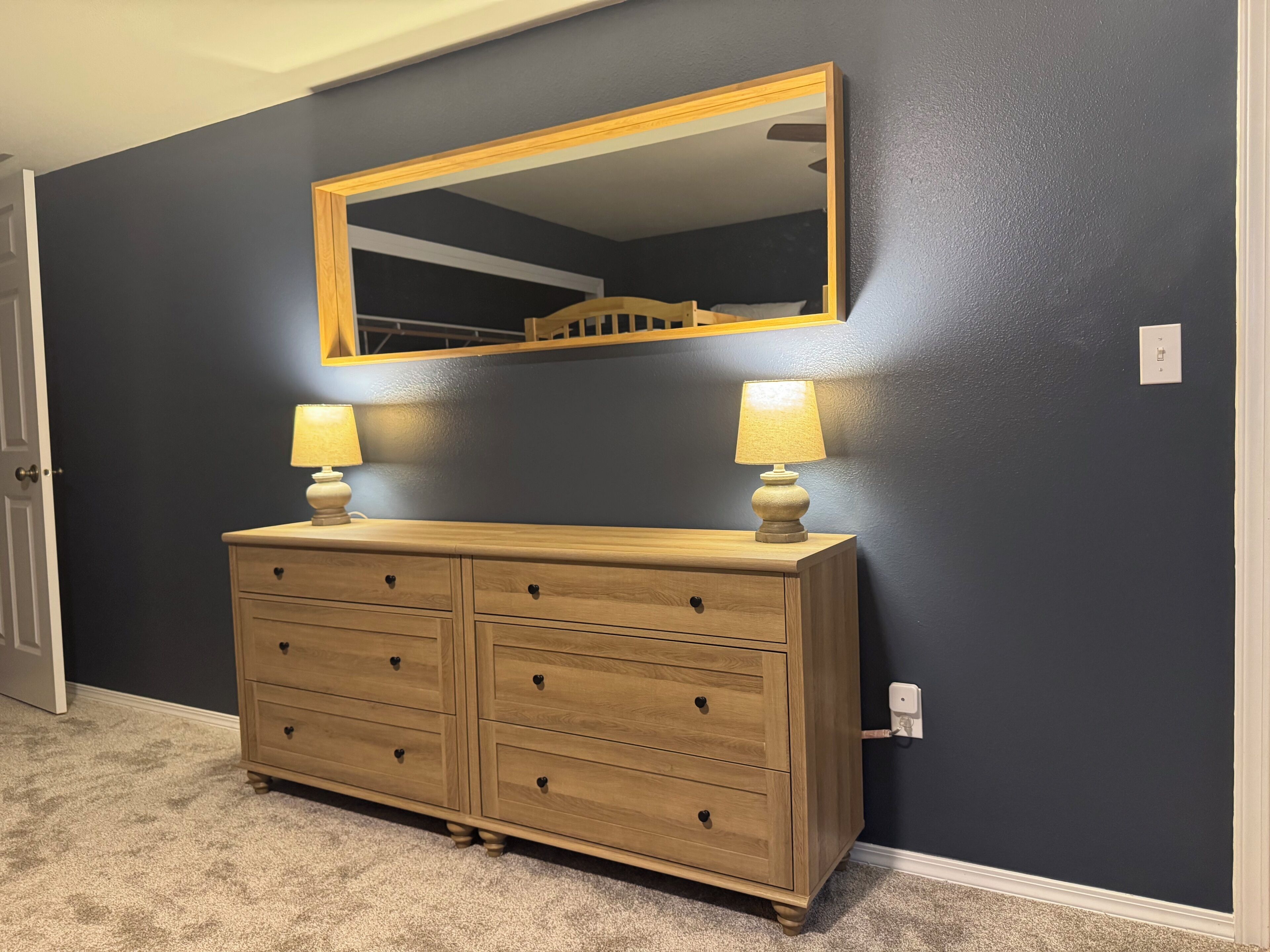 A room with a dark blue wall, a wooden dresser with drawers, two lamps, and a mirror above the dresser.
