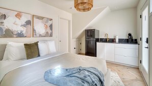 A modern bedroom with a large bed, a kitchen area with white cabinets, and a dining table.