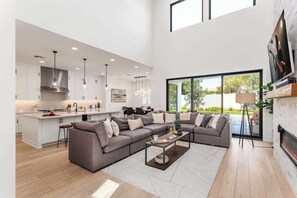 A spacious living area with a large sectional sofa, a coffee table, and a fireplace. There is a kitchen area with white cabinets and pendant lighting.