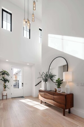 A spacious room with a high ceiling, a wooden console table, a large mirror, and potted plants.