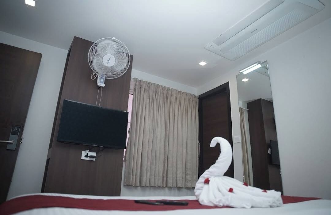 A hotel room with a bed, a wall-mounted TV, a standing fan, a mirror, and a curtain.