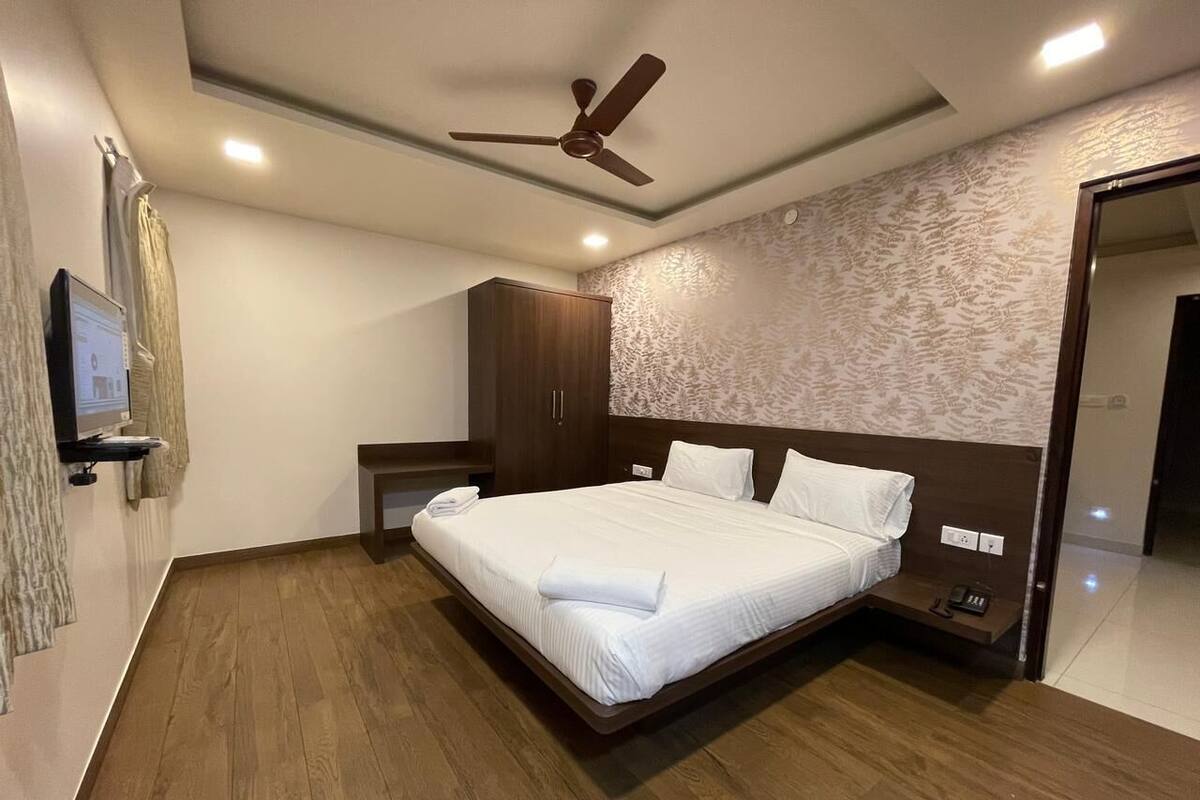 A modern hotel room with a large bed, wooden flooring, a ceiling fan, and a wall-mounted TV.