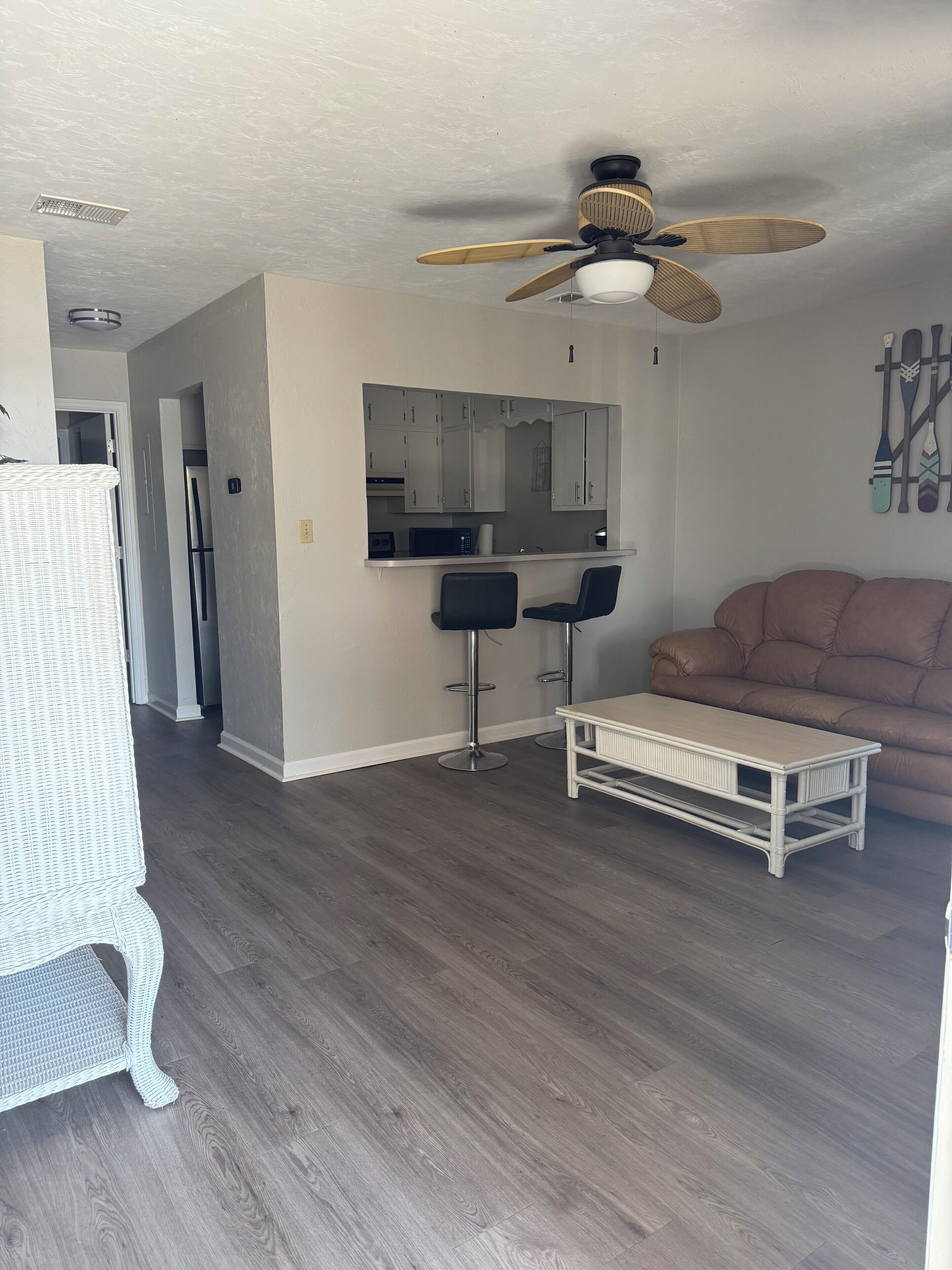 A modern living room with a brown leather sofa, a white coffee table, and two black bar stools. A ceiling fan is visible above the kitchen area, which features white cabinets and a microwave.