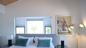 A neatly made bed with white linens and teal pillows, a bedside table with a lamp, and a painting on the wall.