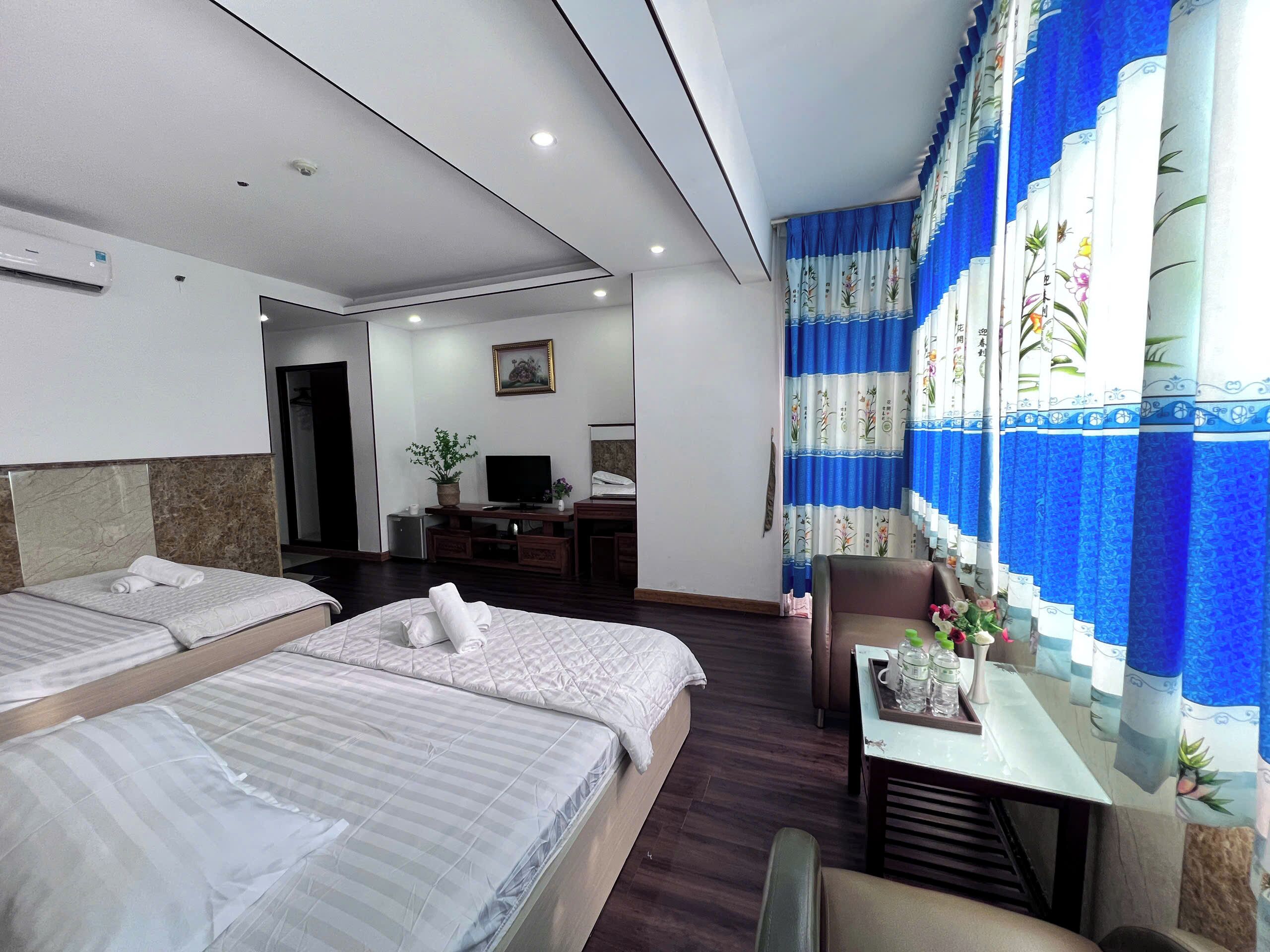 A hotel room with two beds, a desk with a computer, a small TV, and a window with blue curtains.