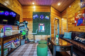 Un intérieur en bois avec un panneau lumineux au néon indiquant l’emplacement de la salle de jeux, un mur vert orné d’un panneau lumineux au néon représentant une manette de jeu, un canapé et une table basse.