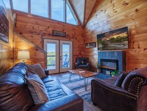 L’intérieur chaleureux d’un chalet avec un canapé en cuir, une petite table, une cheminée et une vue à travers les portes vitrées.