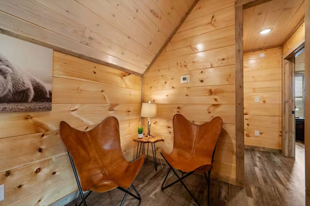 A cozy cabin interior with wooden walls, two leather chairs, a small table with a lamp, and a framed picture on the wall.