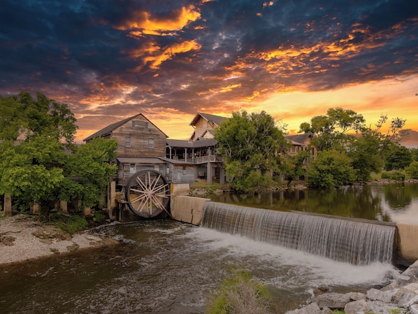 A historic building with a water wheel, a river, and a sunset sky.