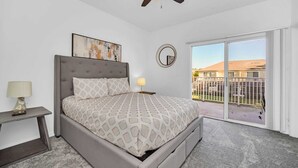 A bedroom with a bed, a nightstand, a ceiling fan, a window with a view of buildings, and a wall-mounted mirror. - Charming 3-bedroom townhome near to attractions (Clermont)