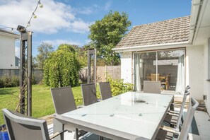 A patio with a glass table and chairs, surrounded by a garden and a house with a tiled roof.