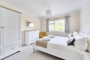 A bedroom with a large wardrobe, a bedside table, a bed with pillows and a blanket, a chair, and a window with curtains.