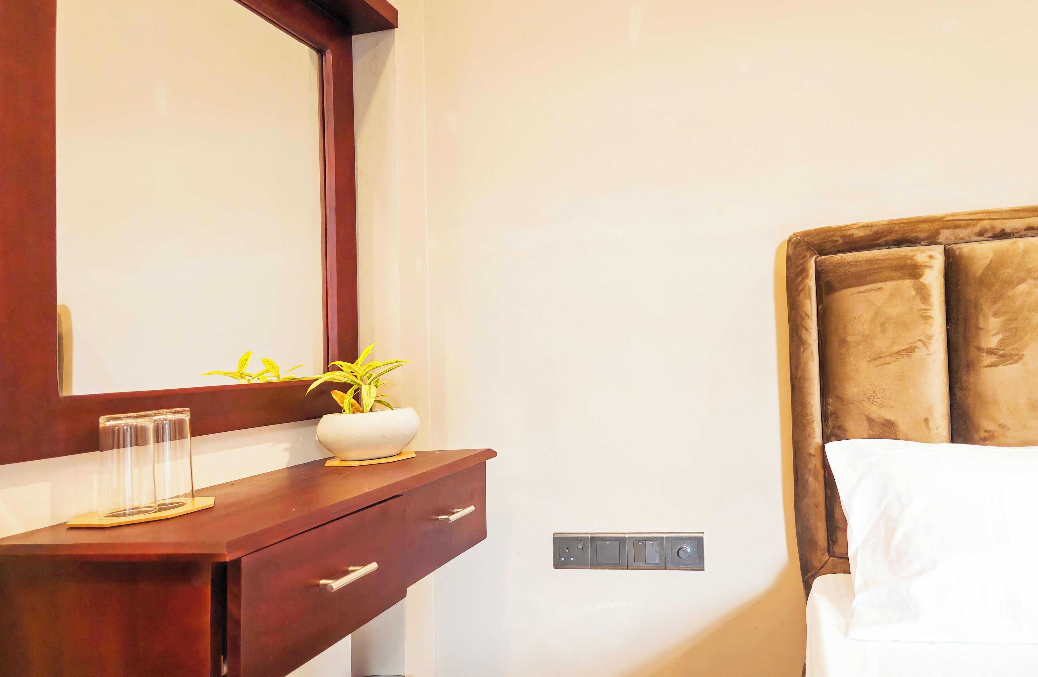 Une chambre d’hôtel avec une coiffeuse en bois, un miroir, une plante en pot, une table de chevet et un lit avec une tête de lit.