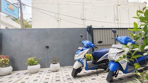 Une zone extérieure dallée avec deux scooters bleus garés, des plantes en pot et un bâtiment en arrière-plan.