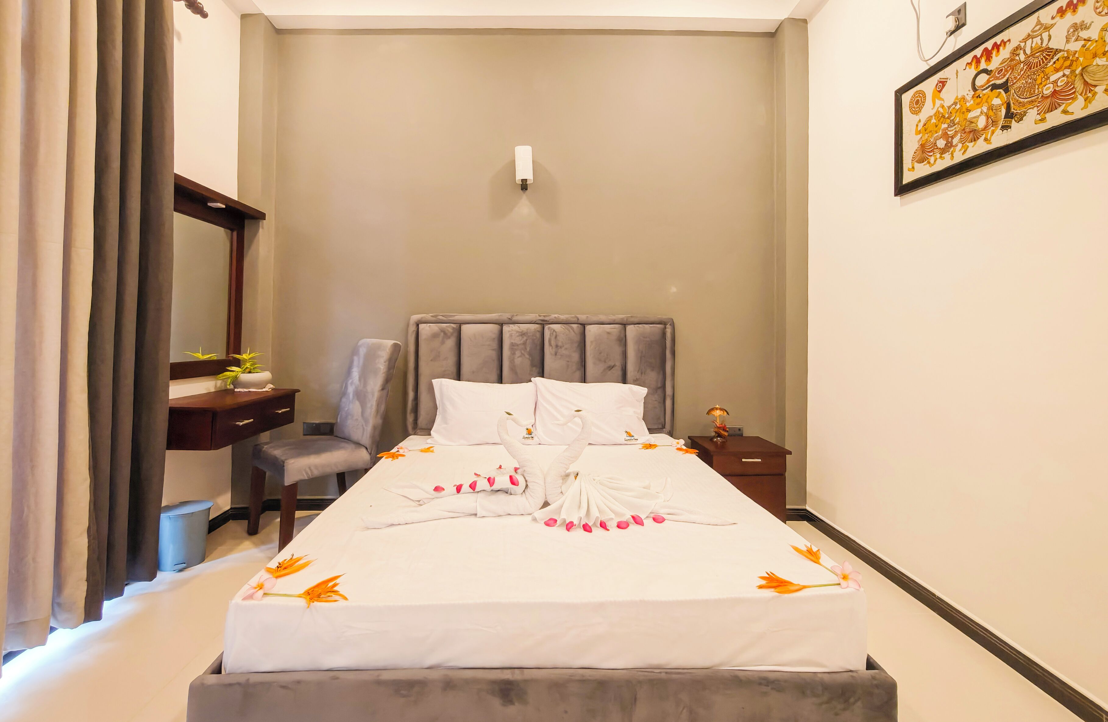 A hotel room with a bed, a desk, a chair, a nightstand, and a wall-mounted light.