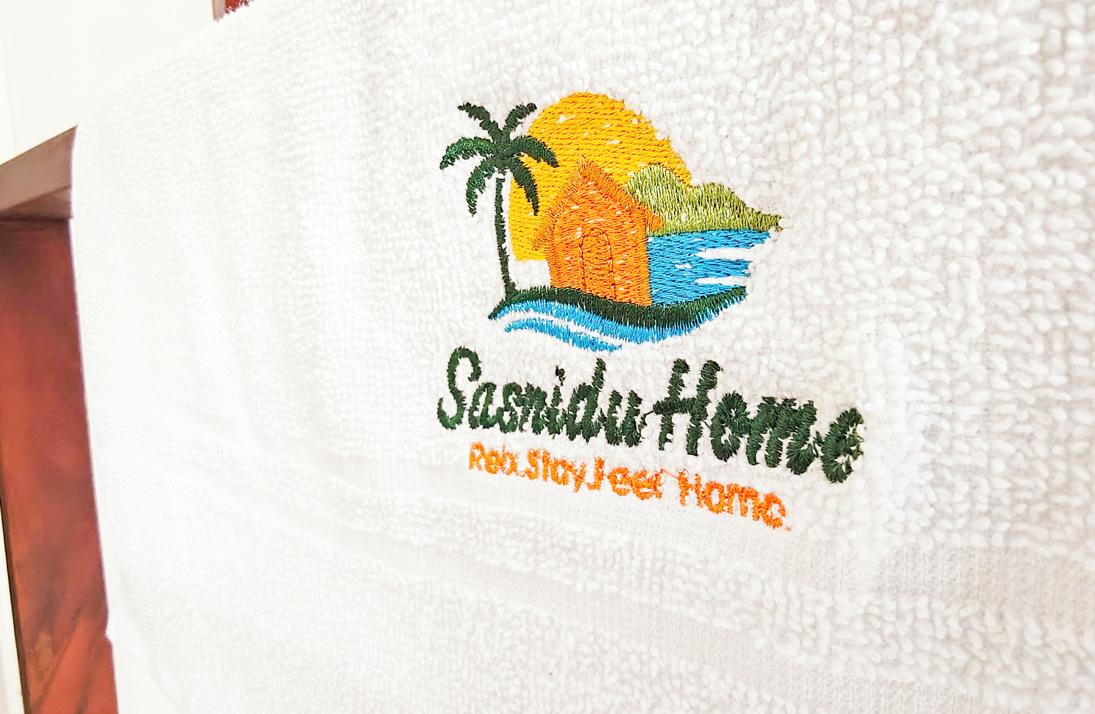 A towel with an embroidered logo and text reading "Sandiut House" and "Enjoy Stay Here Home."