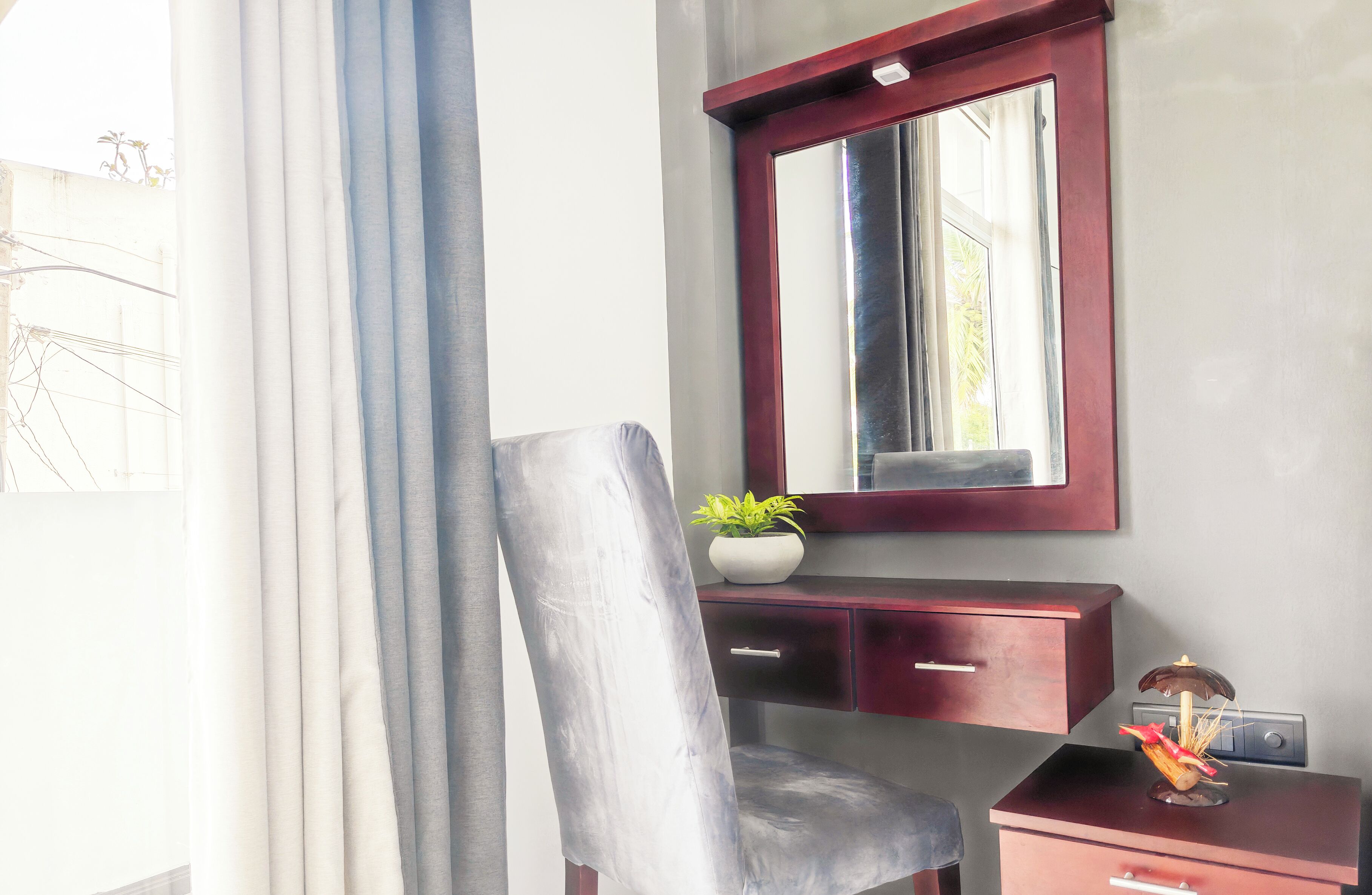 A room with a large mirror, a desk with drawers, a chair, and a small table with a plant and a decorative item.