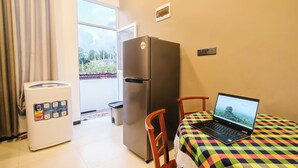 Une petite pièce comprenant un réfrigérateur, un lave-linge, un ordinateur portable posé sur un lit, et une vue à travers une porte.