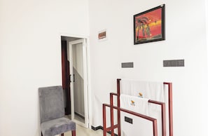 Um quarto com uma cadeira cinza, uma cadeira de madeira com uma toalha, um quadro e uma porta que dá para outro quarto.