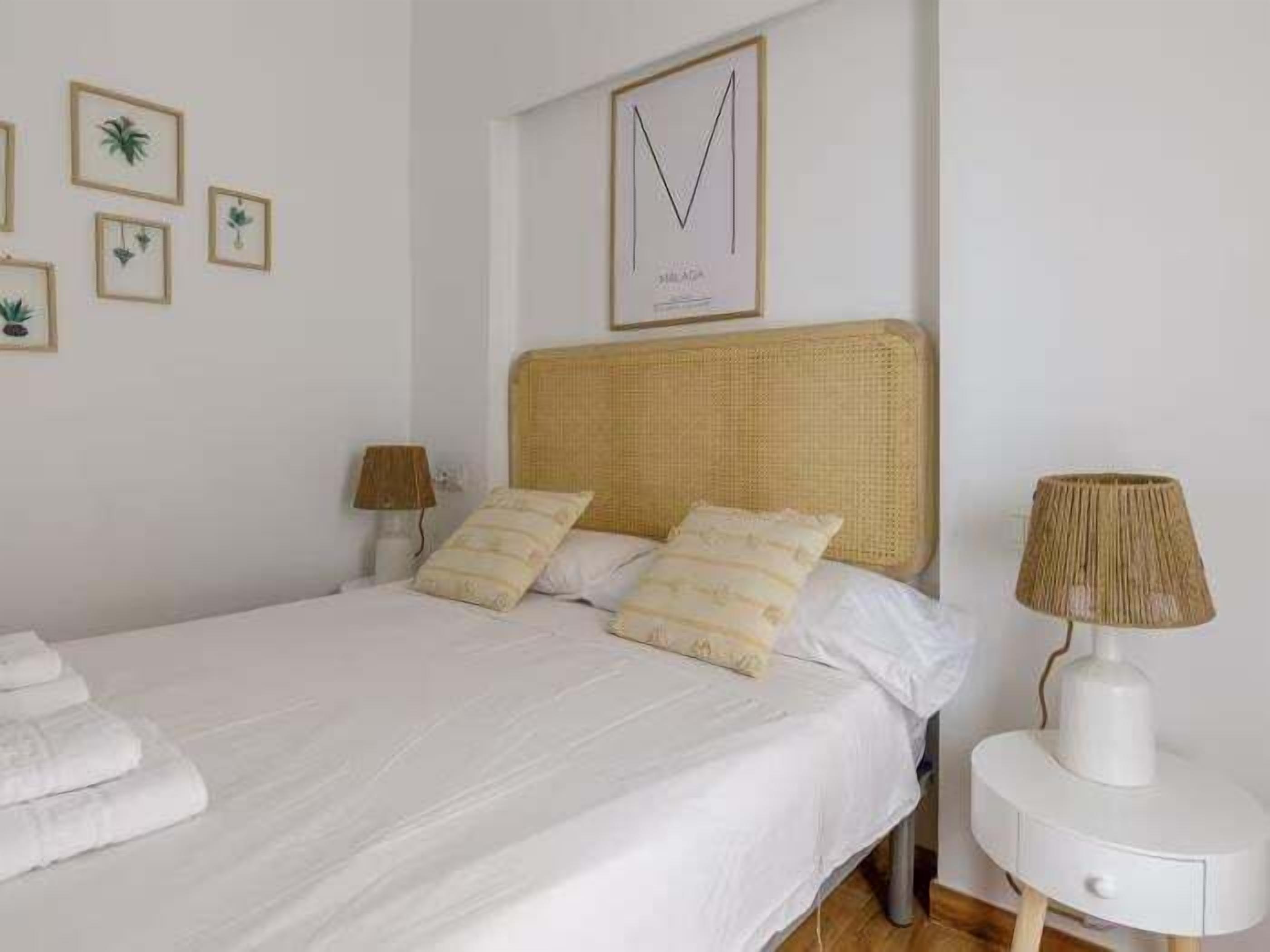 Een net opgemaakt bed met witte lakens en twee kussens, een nachtkastje met een lamp, en wanddecoratie waaronder planten in lijsten en een geometrisch ontwerp.