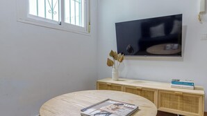 Une pièce avec un téléviseur fixé au mur, une table ronde en bois sur laquelle sont posés des magazines, une console en osier et une fenêtre avec des barreaux en métal.