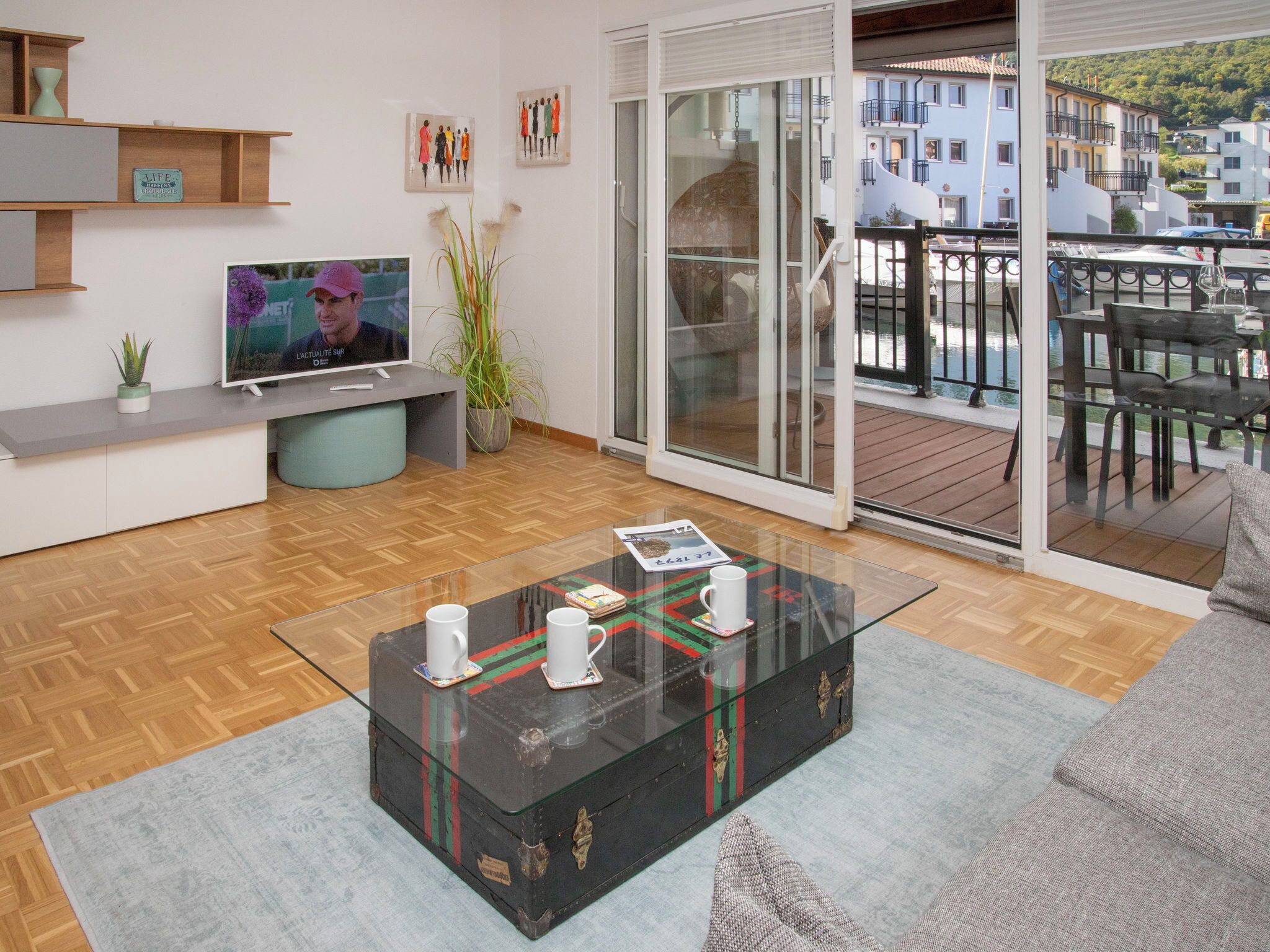 A modern living room with a glass coffee table, a flat-screen TV, and a balcony with outdoor seating.