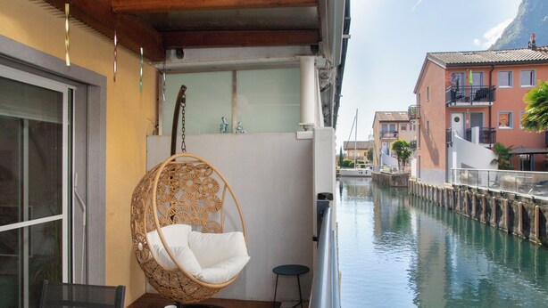 A balcony with a wicker swing chair, a small table with a cup, and a view of a canal with buildings in the background. - Tasman S12/R by Interhome (Le Bouveret)